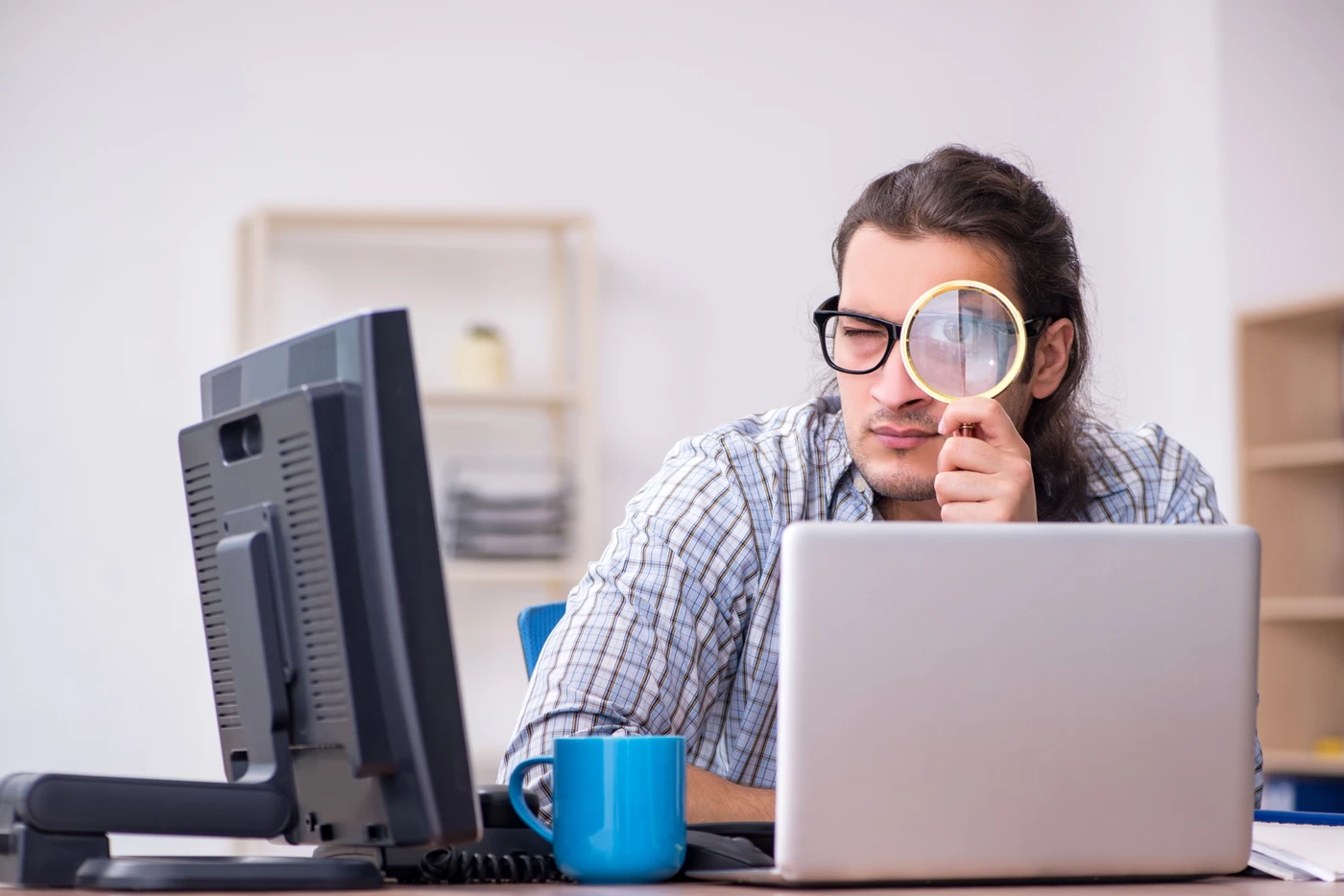 Mężczyzna w okularach patrzy przez lupę na ekran laptopa w biurze – audyt bezpieczeństwa IT i szukanie luk w systemie.
