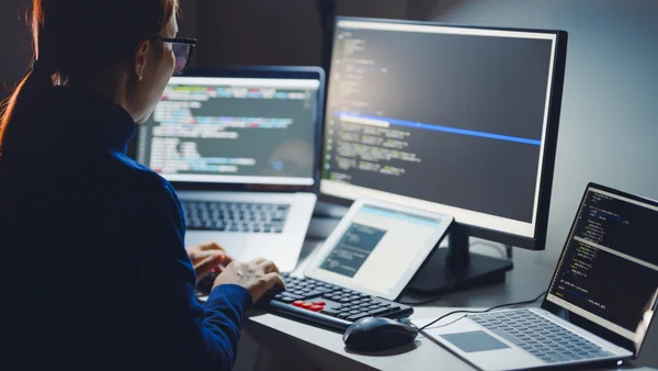 Programistka pracująca przy wielu monitorach z kodem – techniczne wdrażanie systemów bezpieczeństwa i backupu danych.