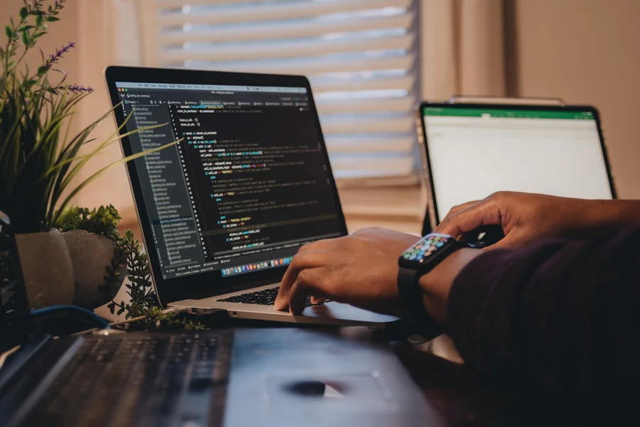 Programista pracujący na laptopie nad zaawansowanym kodem źródłowym, obok widoczny tablet – symbol wiedzy i specjalistycznych usług IT.