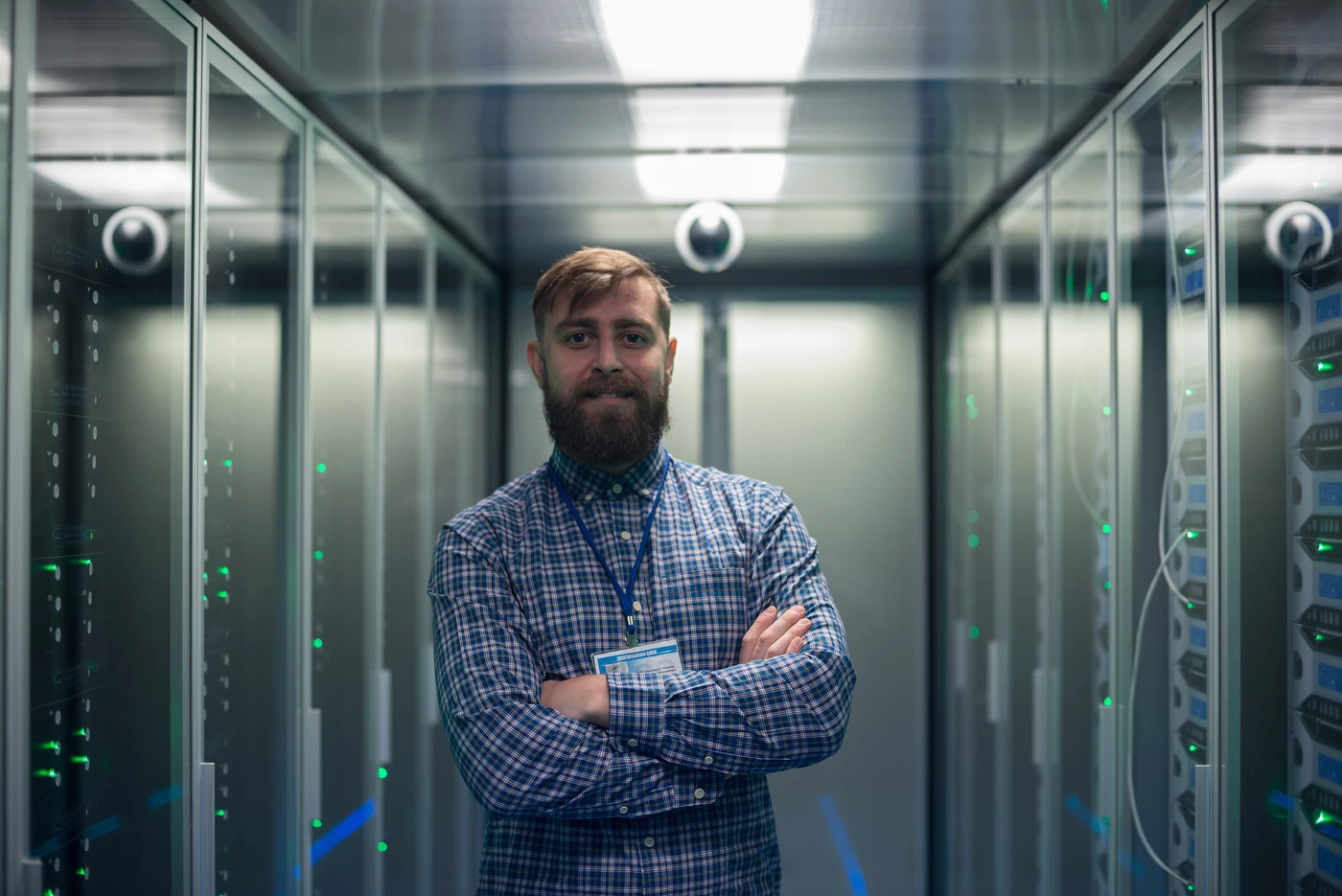 Brodaty mężczyzna w koszuli w kratę z identyfikatorem stoi ze skrzyżowanymi rękami w serwerowni – profesjonalna obsługa informatyczna i monitoring Zabbix.