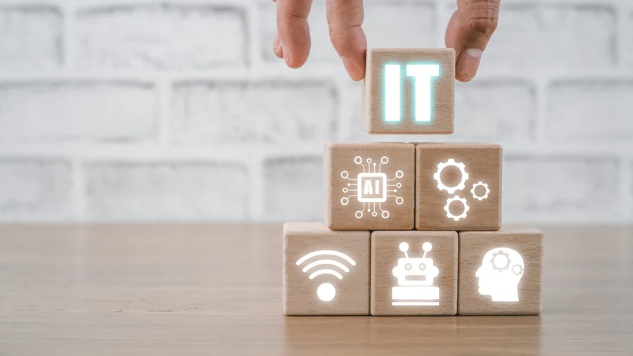 Ręka układająca piramidę z drewnianych klocków z ikonami technologii. Na szczycie świecący klocek z napisem IT, poniżej ikony AI, automatyzacji, sieci i robotyki. Fundamenty nowoczesnego biznesu.