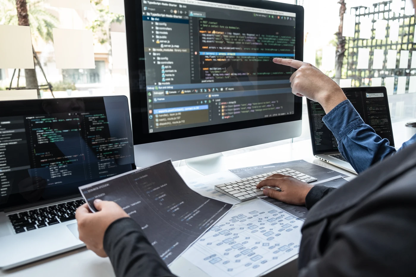 Zbliżenie na biurko programistów Ebimedia analizujących kod w języku TypeScript na dużym monitorze w jasnym, przeszklonym biurze. Dwie osoby współpracują: jedna wskazuje linię kodu na ekranie, druga trzyma wydruk z diagramami technicznymi. Na biurku widać klawiaturę, laptopy i notatki. Zdjęcie w stylu nowoczesnym, dokumentującym pracę specjalistów IT nad integracją systemów lub AI.